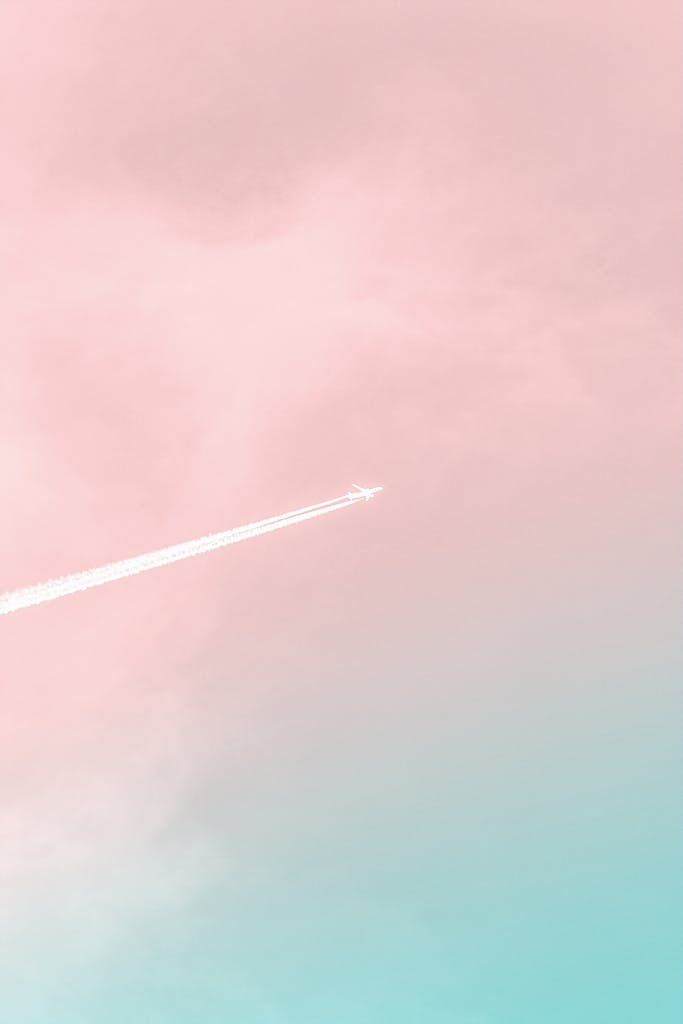 A serene view of an airplane soaring through a pastel pink and blue sky, leaving a contrail.